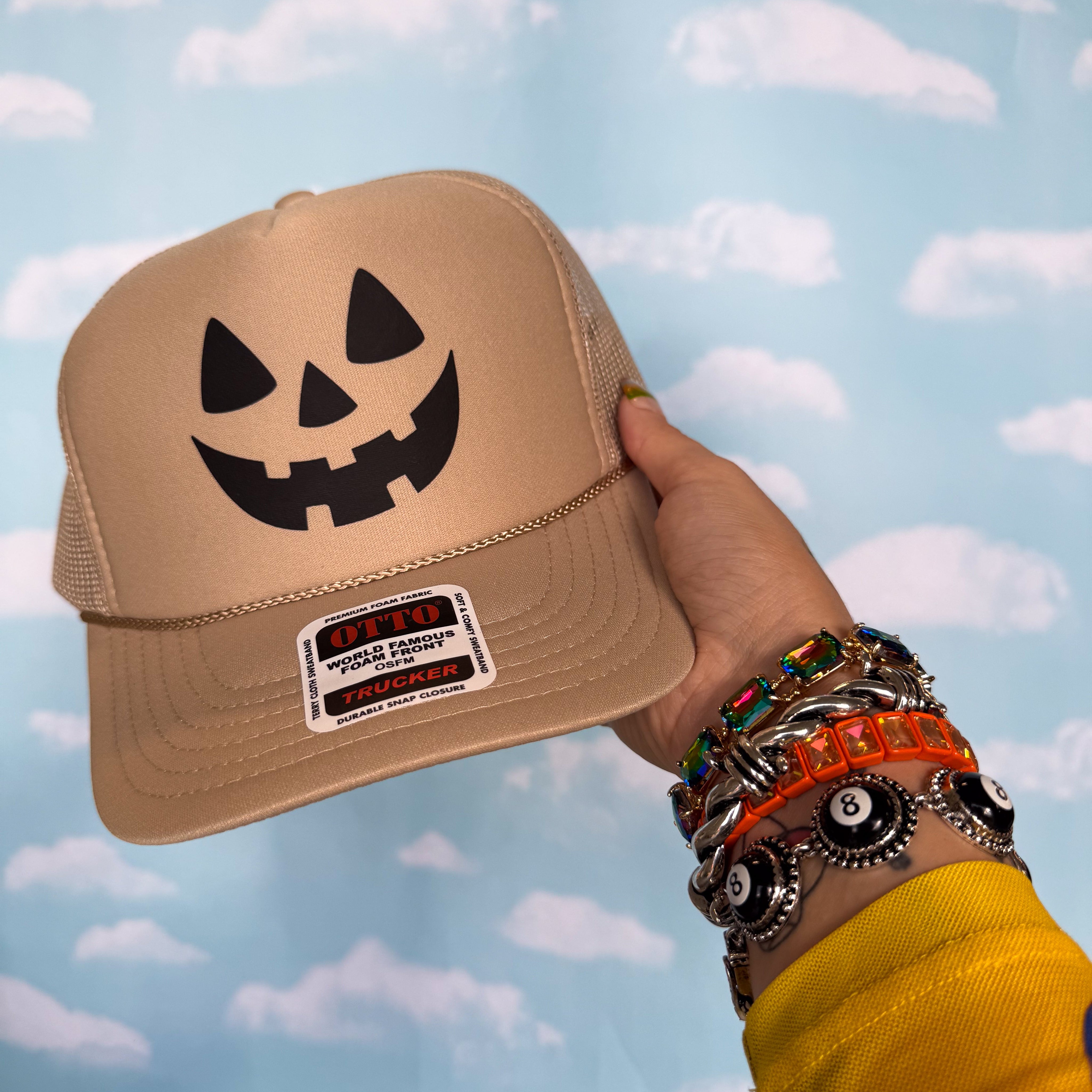 A tan trucker hat with a black jack-o'-lantern face graphic on the front, held by a hand with multiple bracelets against a blue sky and clouds backdrop.