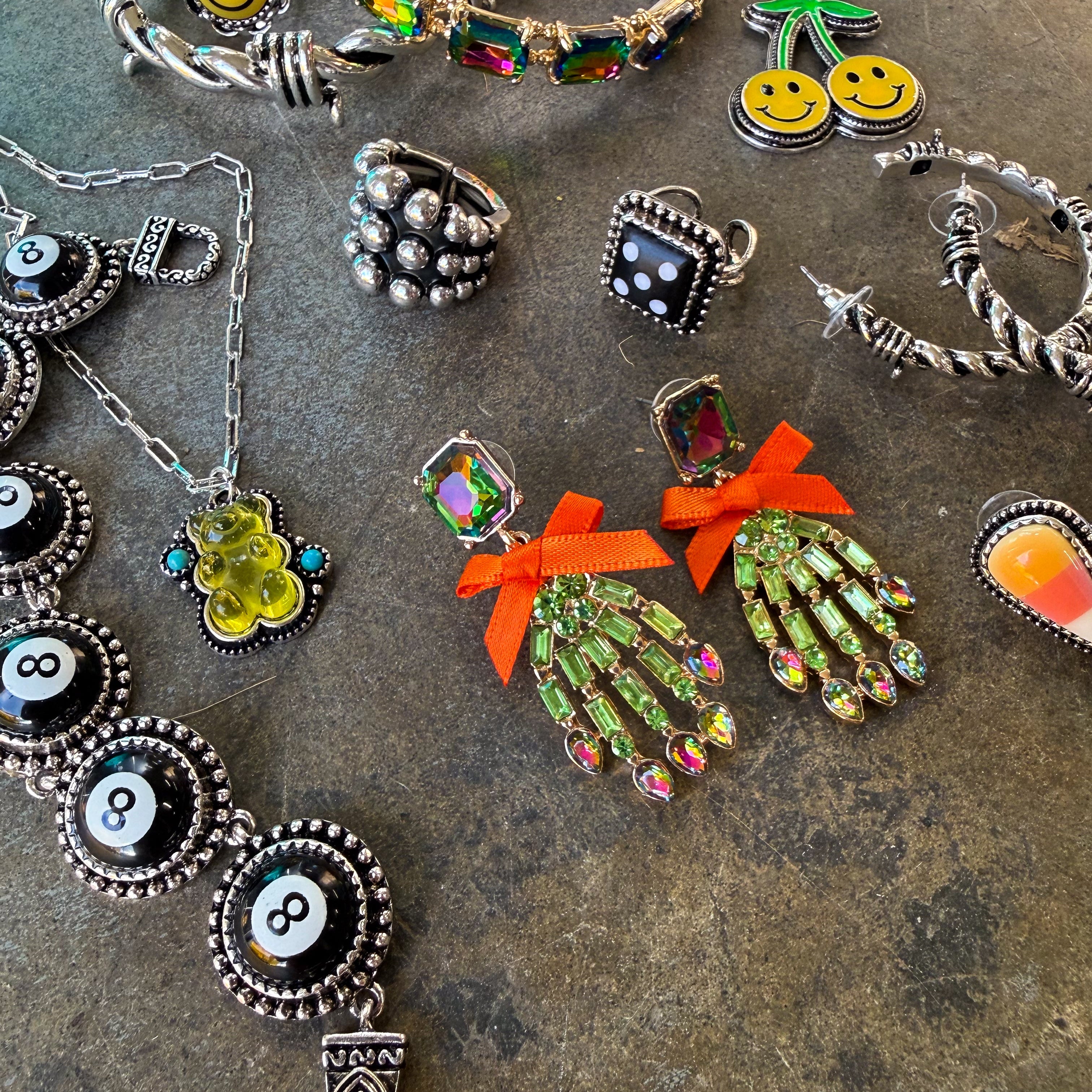 A flat-lay display of various gothic-style jewelry pieces, including silver-toned bracelets, rings, necklaces, and earrings with black, white, green, yellow, orange, and red details, arranged on a textured gray stone surface.