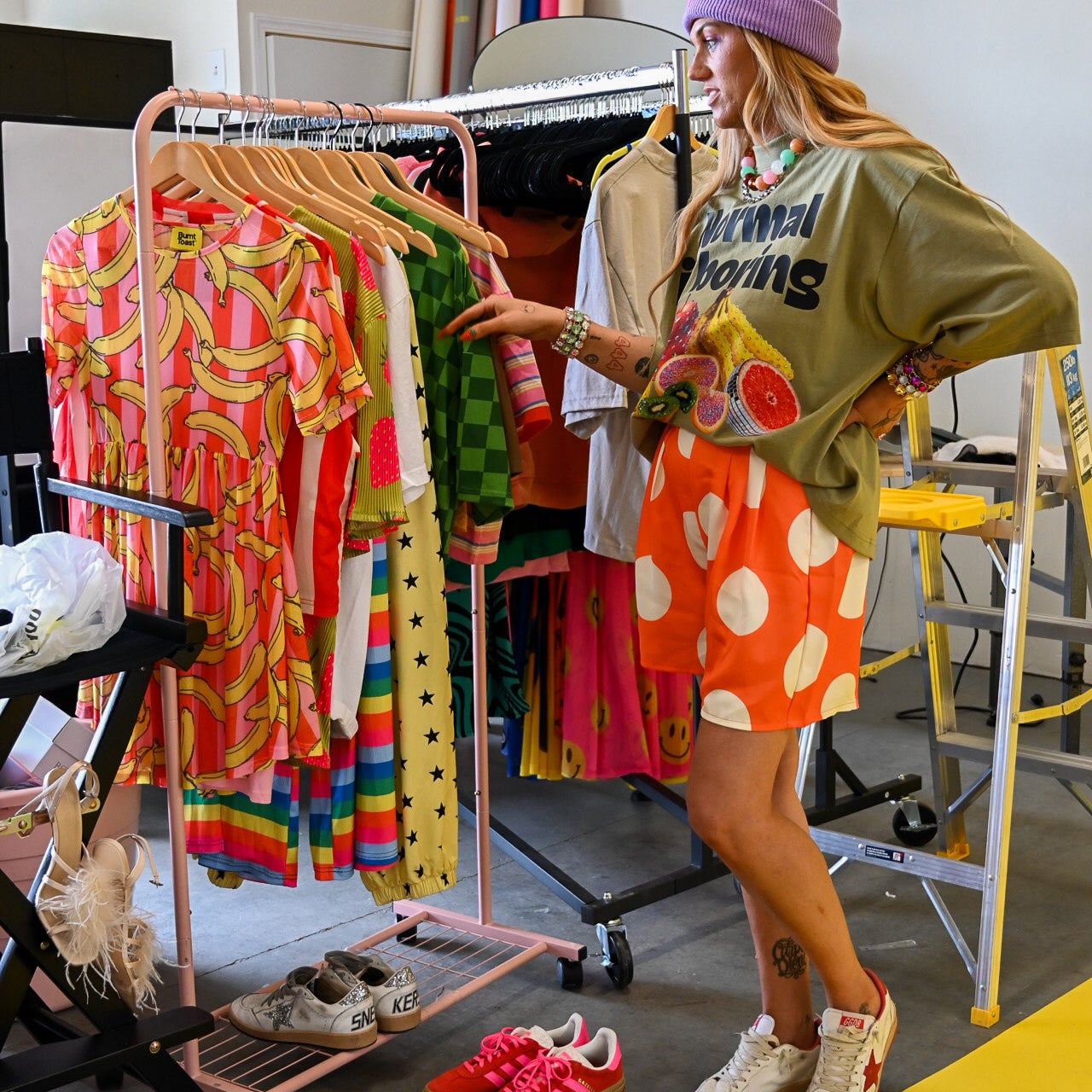 A woman stands in a clothing studio wearing an oversized olive green graphic shirt, a bright orange and white polka dot skirt, white sneakers, and a lavender beanie. She is gesturing toward a pink clothing rack filled with colorful patterned clothing