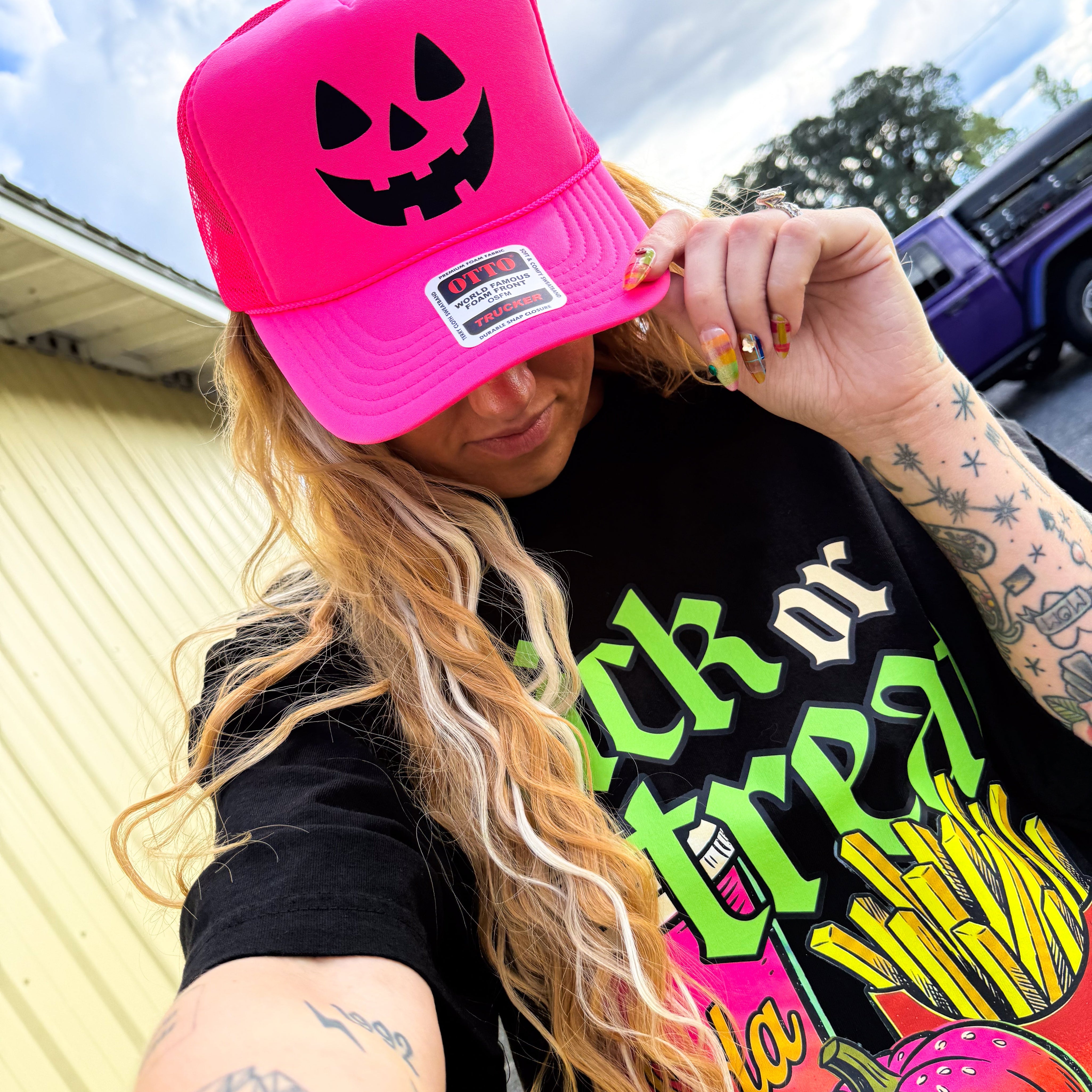 A person wearing a bright pink trucker hat with a black jack-o'-lantern face graphic, paired with a black graphic t-shirt, photographed outdoors in front of a house.