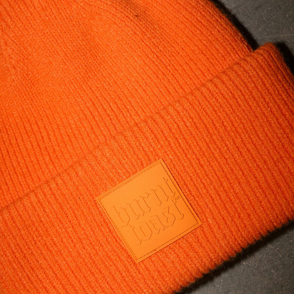 Orange knit beanie with a leather logo patch, on a dark surface, frontal view
