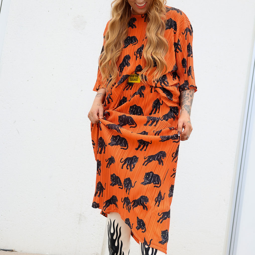 Model wearing an orange plisse short sleeve and skirt set with black animal prints, black beanie, and black and white boots, standing outdoors, frontal view