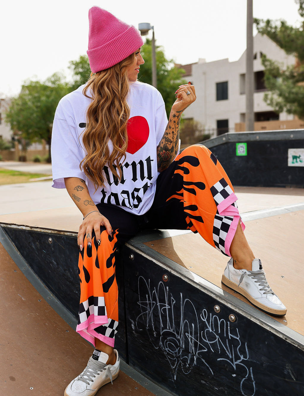 Model wearing a white t-shirt with a red heart, black text, pink beanie, black pants with orange flame, checkered design, and pink hem at the ankle, pink beanie, sitting on a skateboard ramp, frontal view
