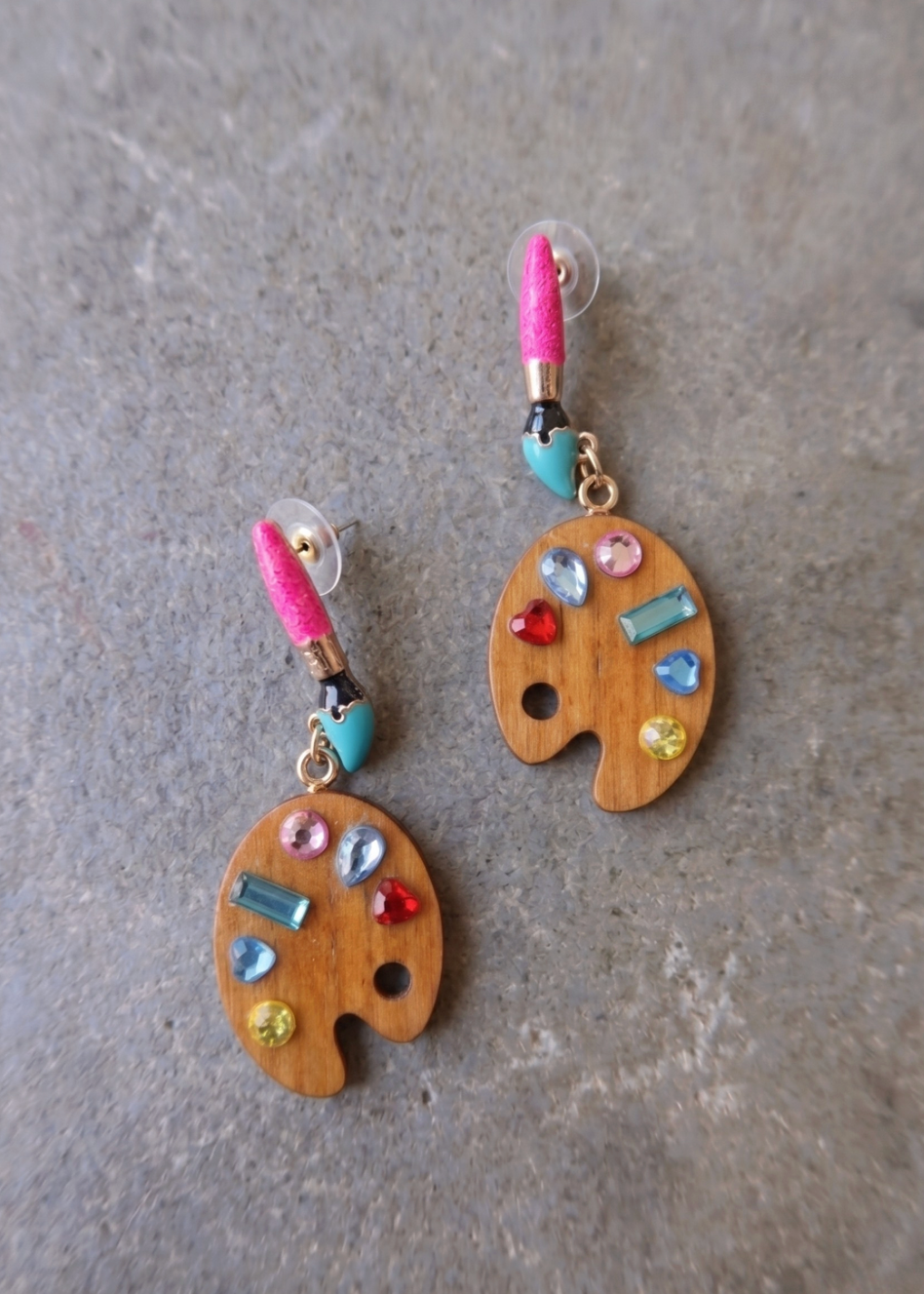 A pair of dangling earrings shaped like wooden paint palettes, decorated with colorful rhinestones and a pink glitter paintbrush, displayed on a textured gray surface