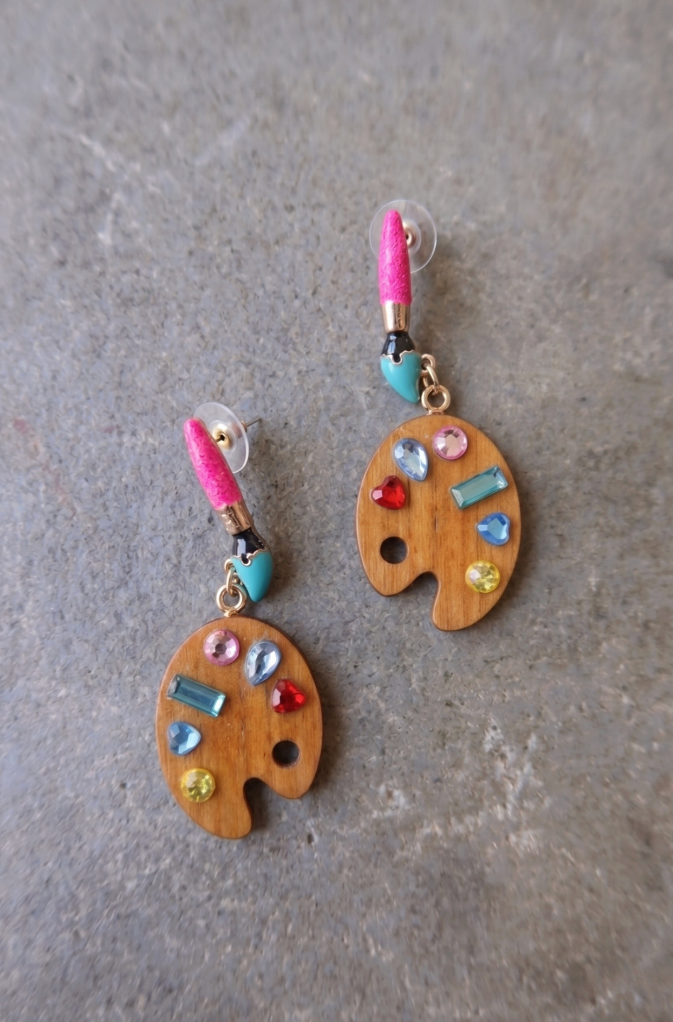 A pair of dangling earrings shaped like wooden paint palettes, decorated with colorful rhinestones and a pink glitter paintbrush, displayed on a textured gray surface