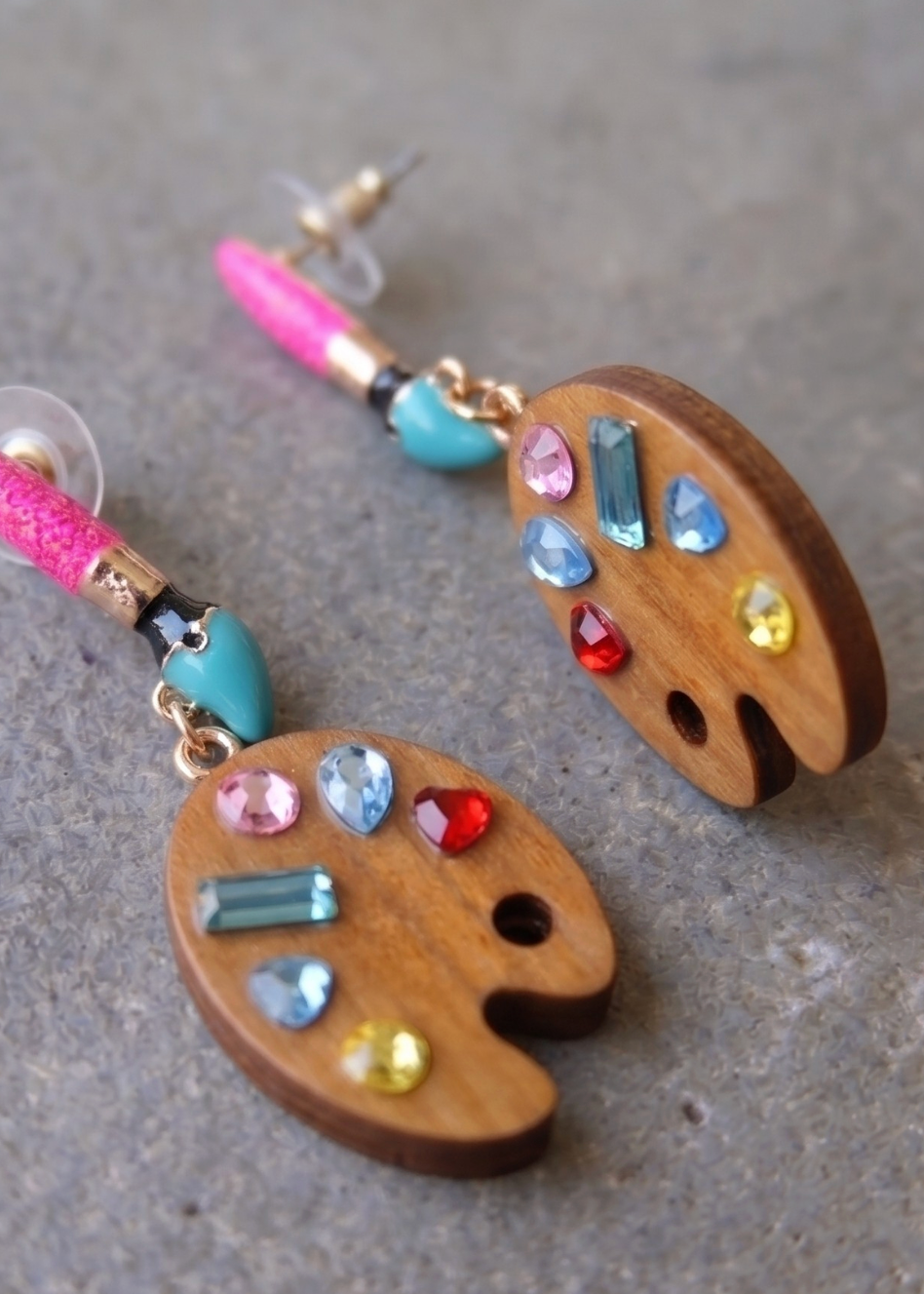 A pair of wooden artist palette drop earrings decorated with colorful gemstones, displayed on a textured gray surface