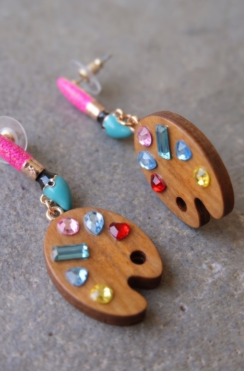 A pair of wooden artist palette drop earrings decorated with colorful gemstones, displayed on a textured gray surface