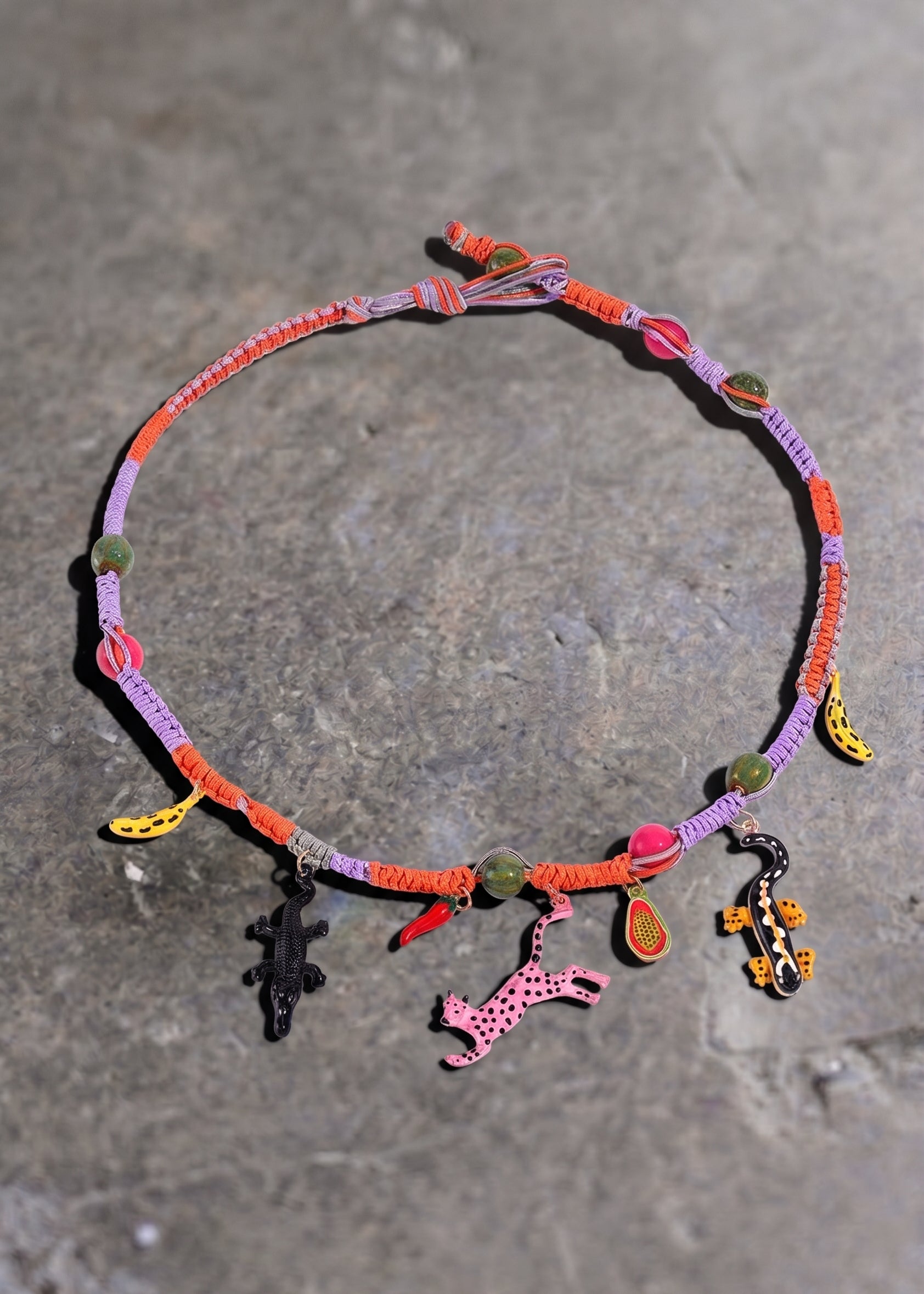 A colorful braided cord necklace with small charms, including a pink giraffe, a black crocodile, and yellow and orange accents, displayed on a gray stone surface
