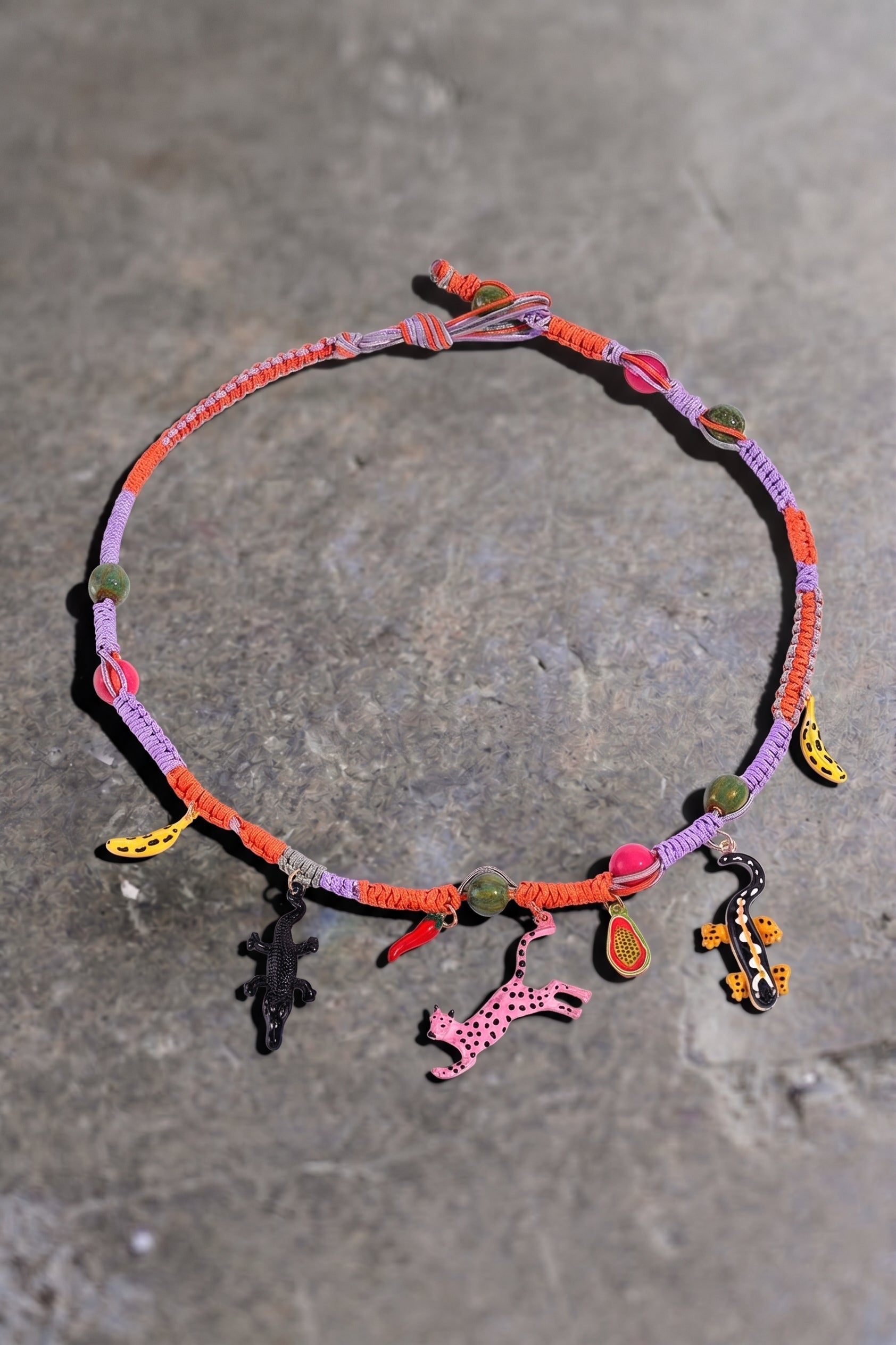 A colorful braided cord necklace with small charms, including a pink giraffe, a black crocodile, and yellow and orange accents, displayed on a gray stone surface