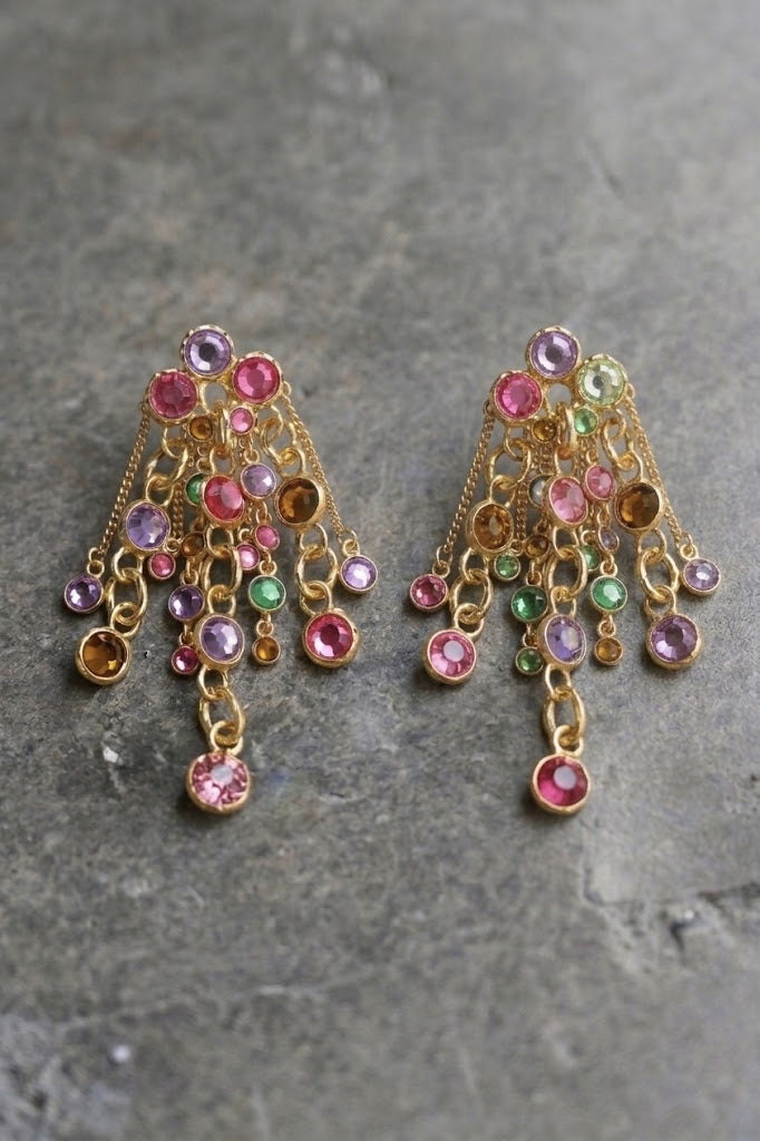 A pair of gold-toned chandelier earrings with dangling chains and multiple round gemstones in pink, purple, green, and amber tones, displayed on a textured gray stone surface