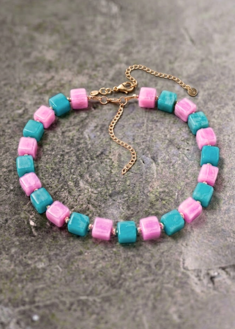 A beaded necklace with alternating pink and turquoise square beads, finished with a gold-tone clasp and extender chain, displayed on a textured gray stone surface
