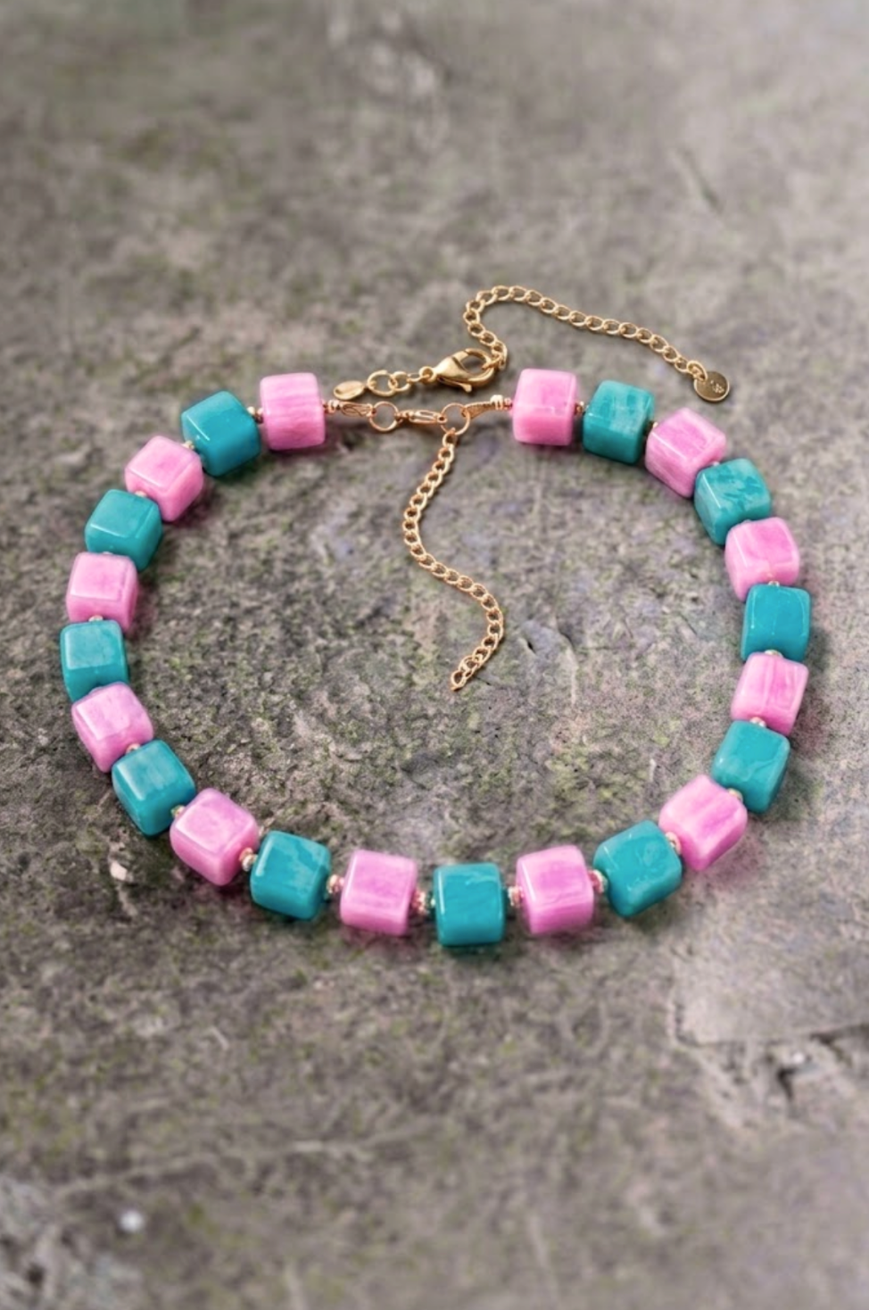 A beaded necklace with alternating pink and turquoise square beads, finished with a gold-tone clasp and extender chain, displayed on a textured gray stone surface