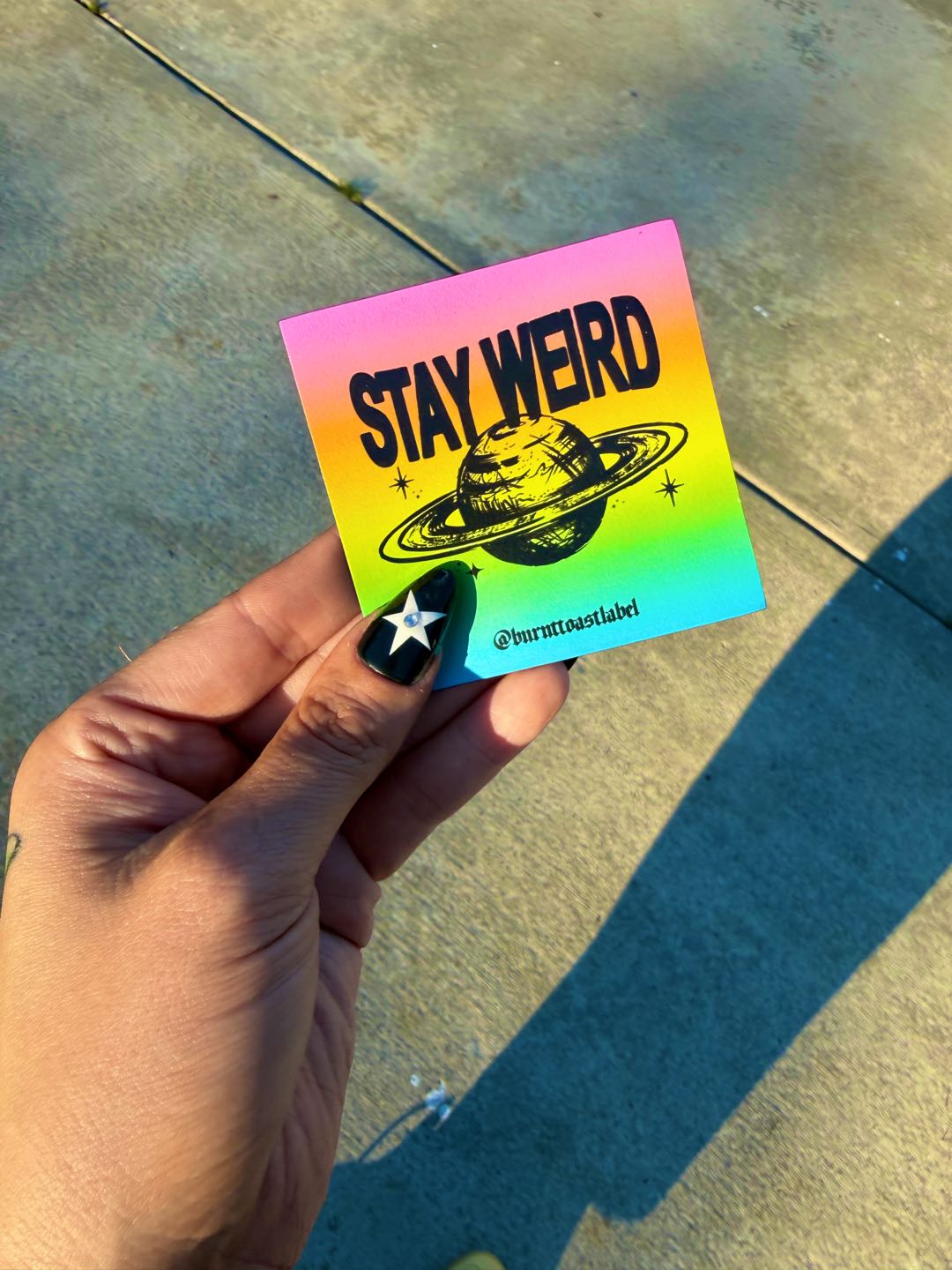 A hand holding a magnet with a rainbow gradient and the words STAY WEIRD with a planet in black, on a concrete background