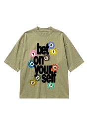 An oversized short-sleeve t-shirt in olive green, featuring a graphic print on the front with the text 'bet on your self' in black and colorful bingo balls with numbers.