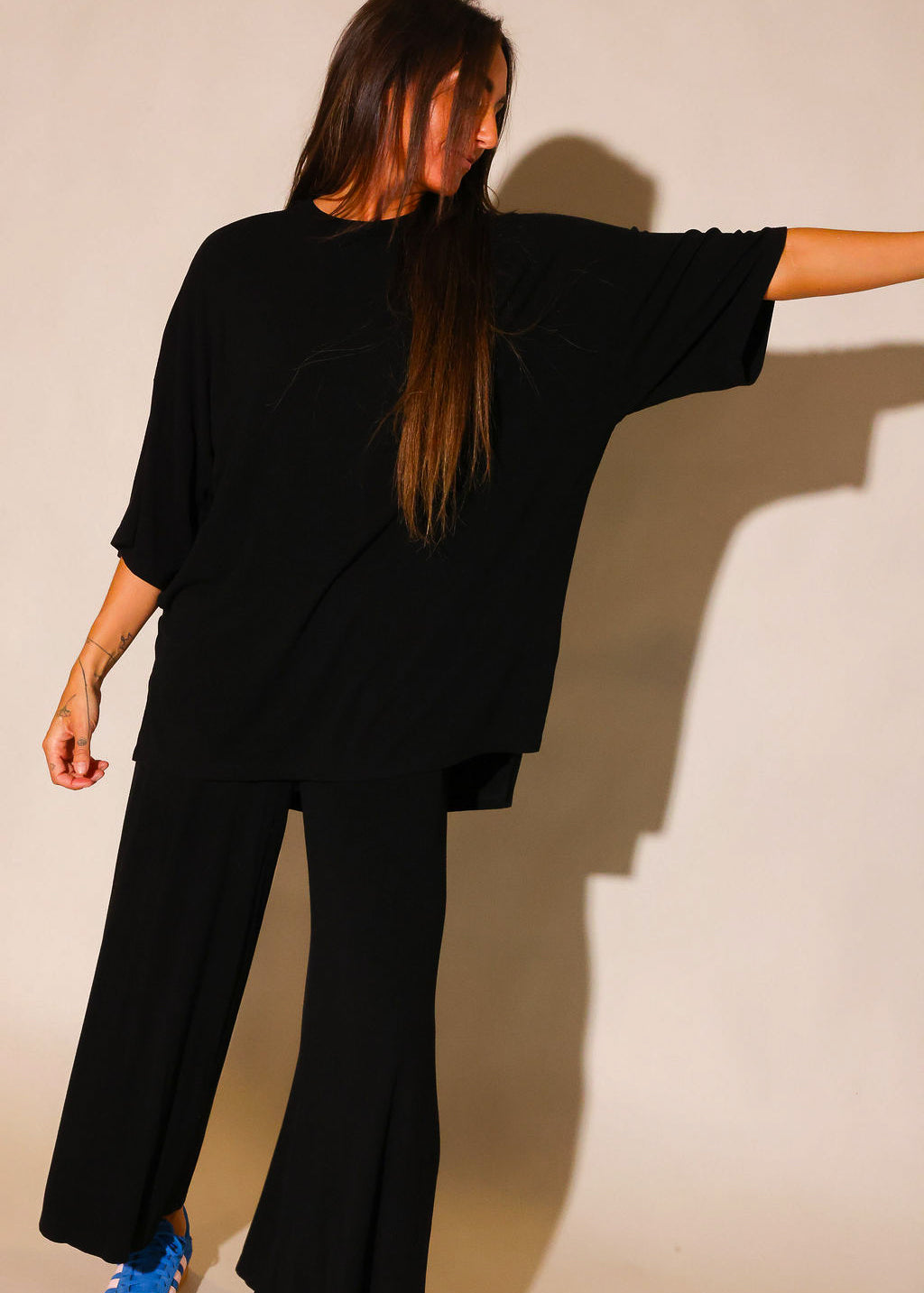 Front view of a model wearing a black ribbed two piece set, including a loose fitting, short sleeve top and high waisted pants. The model is standing with a relaxed posture and wearing blue sneakers