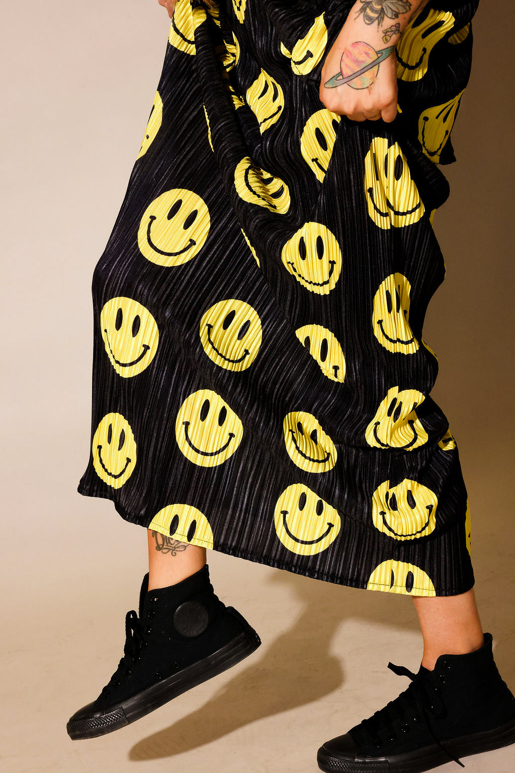 Model wearing a black short sleeve and long skirt plisse set with yellow smiley face patterns, black tennis shoes, standing with one foot lifted, side view