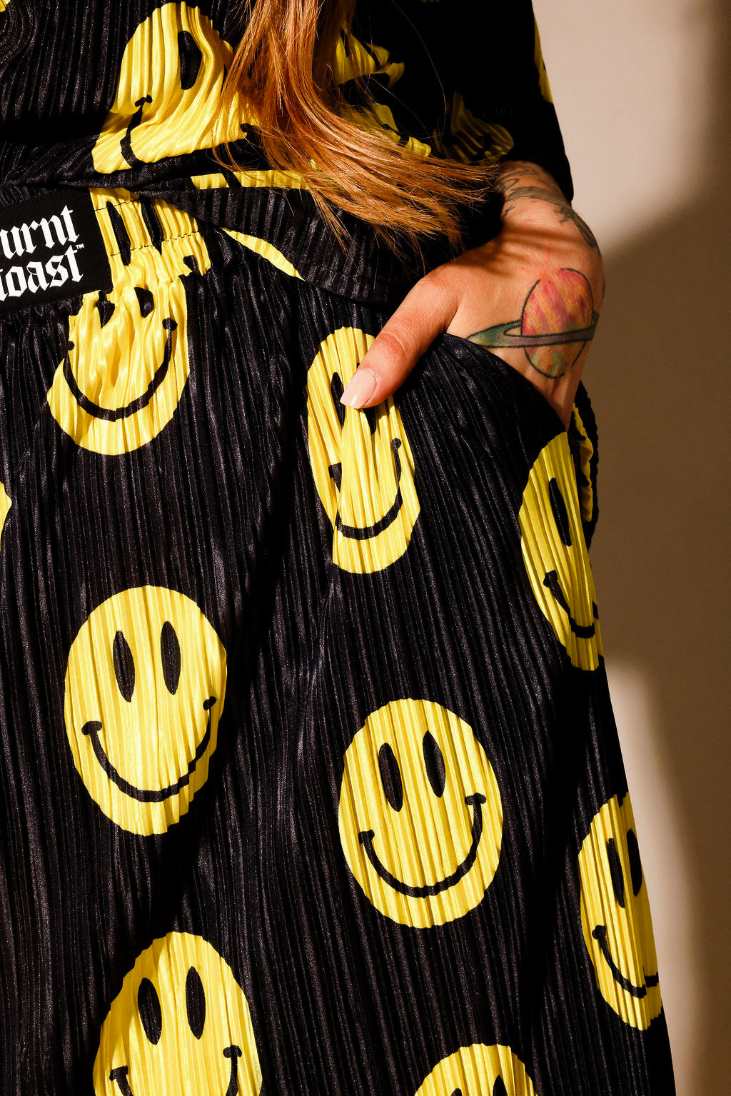 Model wearing a black short sleeve and long skirt plisse set with yellow smiley face patterns, burnt toast label on drawstring waist band, with hand in pocket, standing, close up frontal view