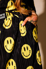 Model wearing a black short sleeve and long skirt plisse set with yellow smiley face patterns, burnt toast label on drawstring waist band, with hand in pocket, standing, close up frontal view