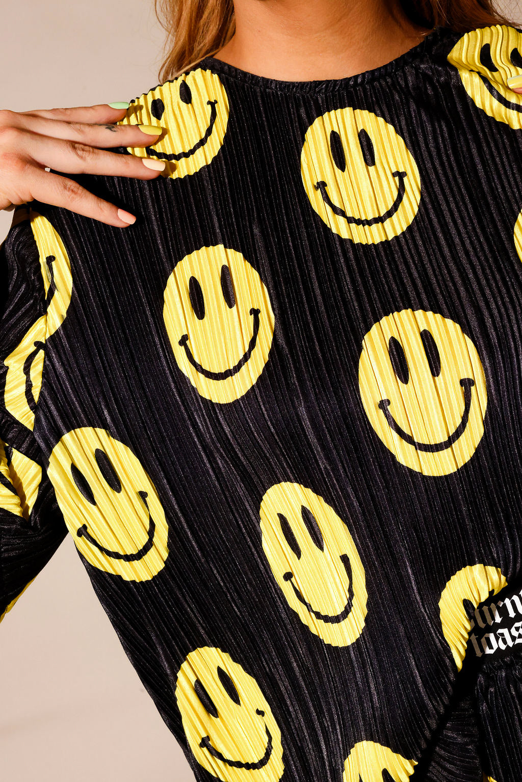 Model wearing a black short sleeve and long skirt plisse set with yellow smiley face patterns, with hand on shoulder, showing neckline, standing, close up frontal view
