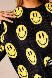 Model wearing a black short sleeve and long skirt plisse set with yellow smiley face patterns, with hand on shoulder, showing neckline, standing, close up frontal view