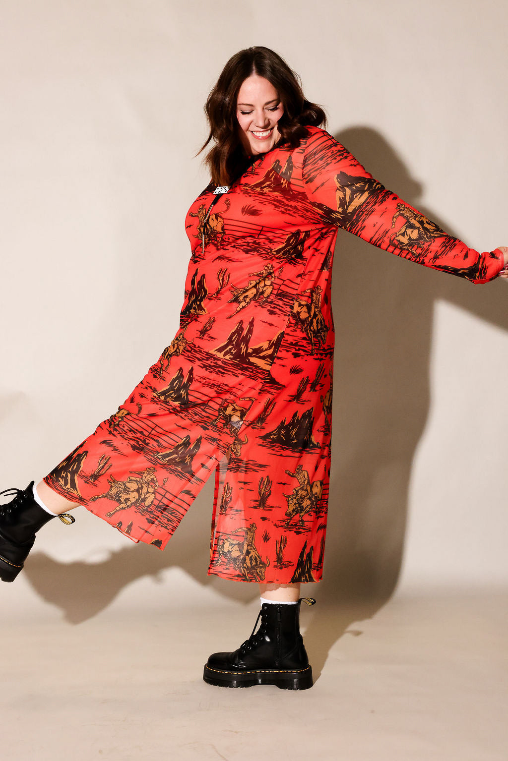 A woman modeling a long-sleeved red midi dress with a black and gold scenic print, paired with black boots, against a plain beige studio background, frontal view