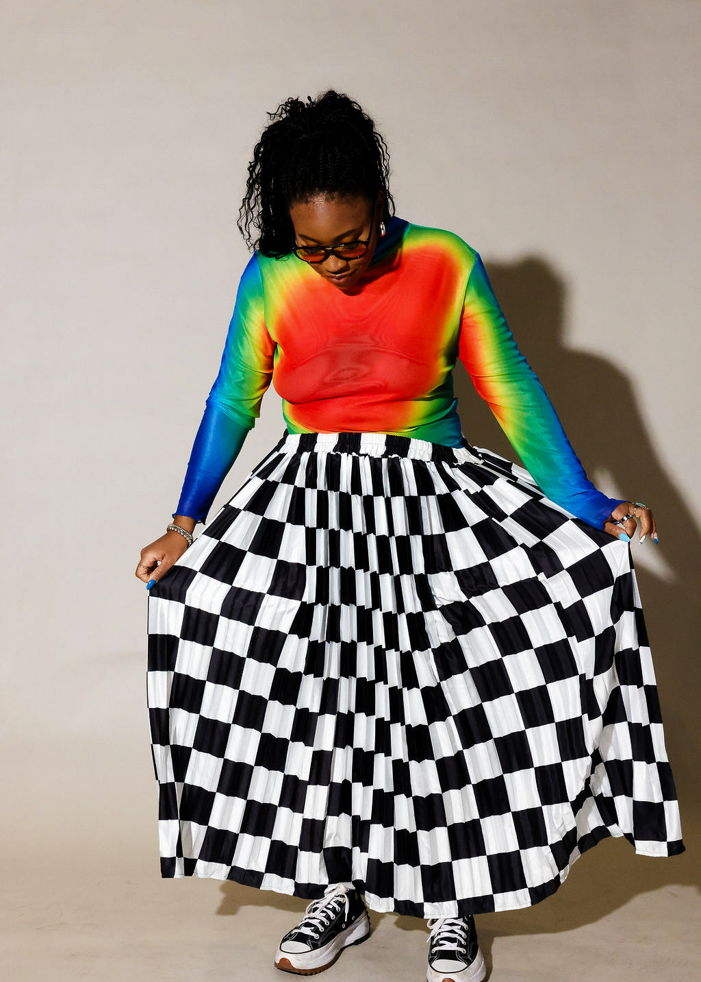 A person wearing a long-sleeve rainbow tie-dye top and a black-and-white checkered pleated skirt, holding out the skirt to show its full volume against a plain beige studio background, frontal view