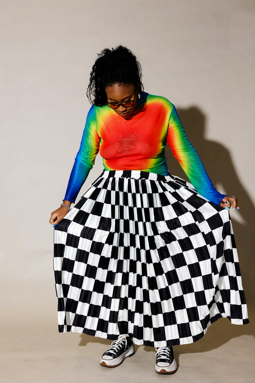 A person wearing a long-sleeve rainbow tie-dye top and a black-and-white checkered pleated skirt, holding out the skirt to show its full volume against a plain beige studio background, frontal view