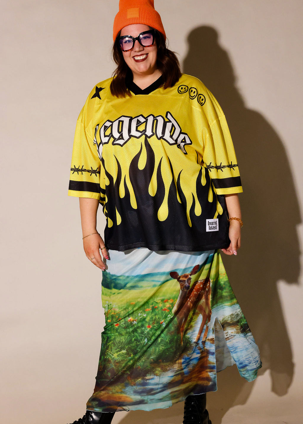 A person modeling a yellow and black graphic jersey with flame print and a long multicolor landscape print skirt, paired with an orange beanie, black sunglasses, and black boots against a plain beige studio background, frontal view