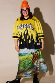 A person modeling a yellow and black graphic jersey with flame print and a long multicolor landscape print skirt, paired with an orange beanie, black sunglasses, and black boots against a plain beige studio background, frontal view