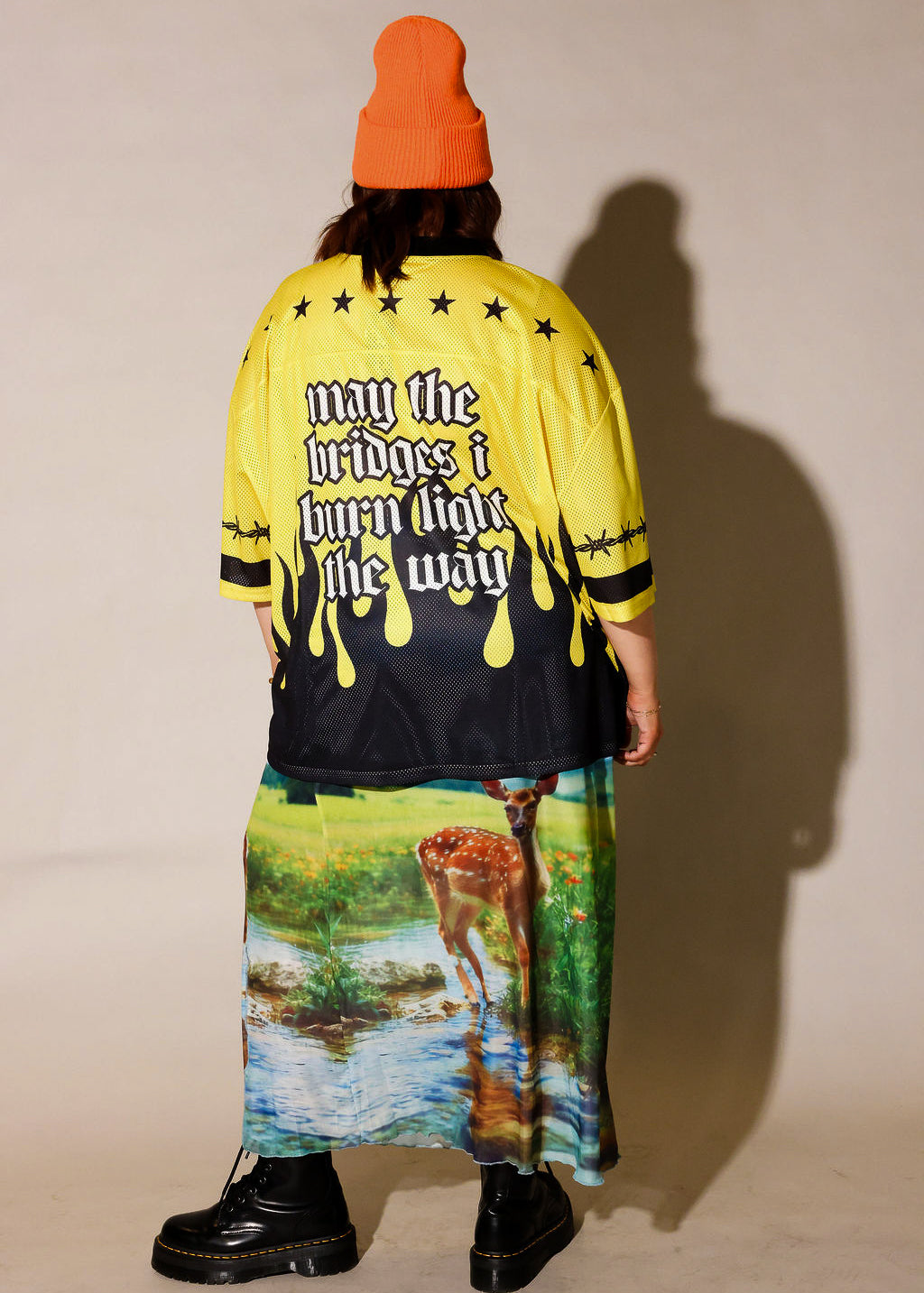 A person shown from the back wearing a bright yellow and black graphic jacket with the phrase 'may the bridges i burn light the way' in white gothic lettering, paired with a long multicolor scenic print skirt and an orange beanie, against a plain beige studio background, back view