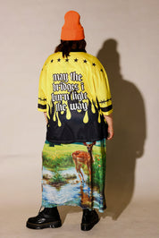 A person shown from the back wearing a bright yellow and black graphic jacket with the phrase 'may the bridges i burn light the way' in white gothic lettering, paired with a long multicolor scenic print skirt and an orange beanie, against a plain beige studio background, back view