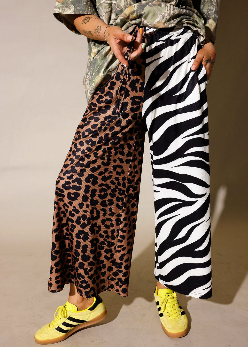 Model is wearing a camo graphic shirt, half leopard print pants, half zebra print pants on a purple background, close up frontal view