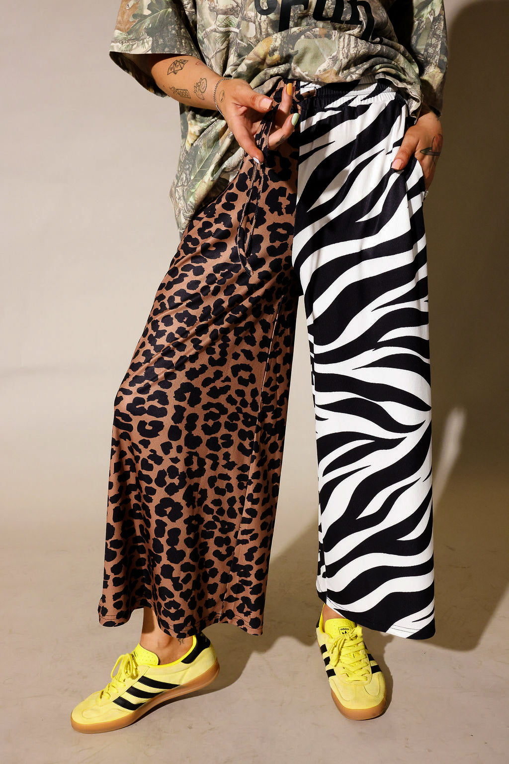 Model is wearing a camo graphic shirt, half leopard print pants, half zebra print pants on a purple background, close up frontal view