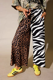 Model is wearing a camo graphic shirt, half leopard print pants, half zebra print pants on a purple background, close up frontal view