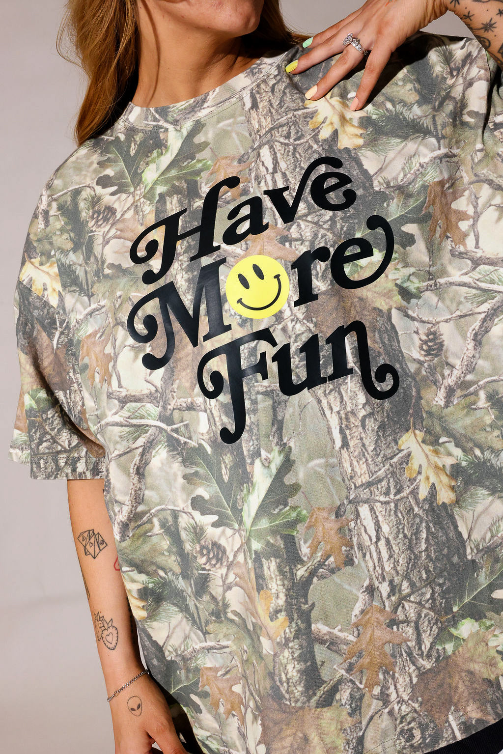 Model wearing a camo graphic tee with 'Have more fun' text on a beige background