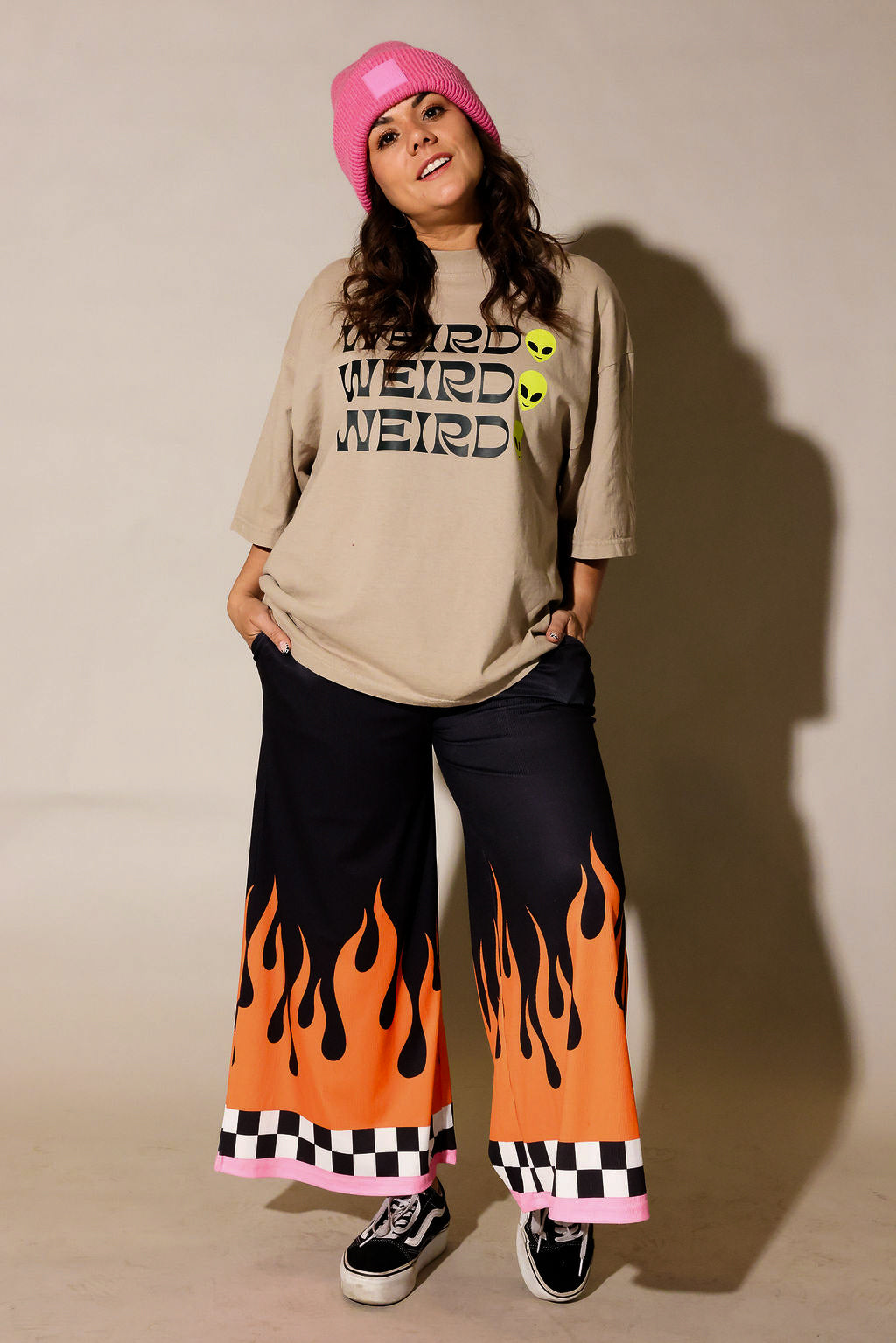 A model wearing a beige graphic t-shirt with 'WEIRD' printed in black and yellow, black wide-leg pants with an orange and black flame pattern, a pink beanie, and black sneakers, standing against a beige wall