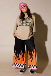 A model wearing a beige graphic t-shirt with 'WEIRD' printed in black and yellow, black wide-leg pants with an orange and black flame pattern, a pink beanie, and black sneakers, standing against a beige wall