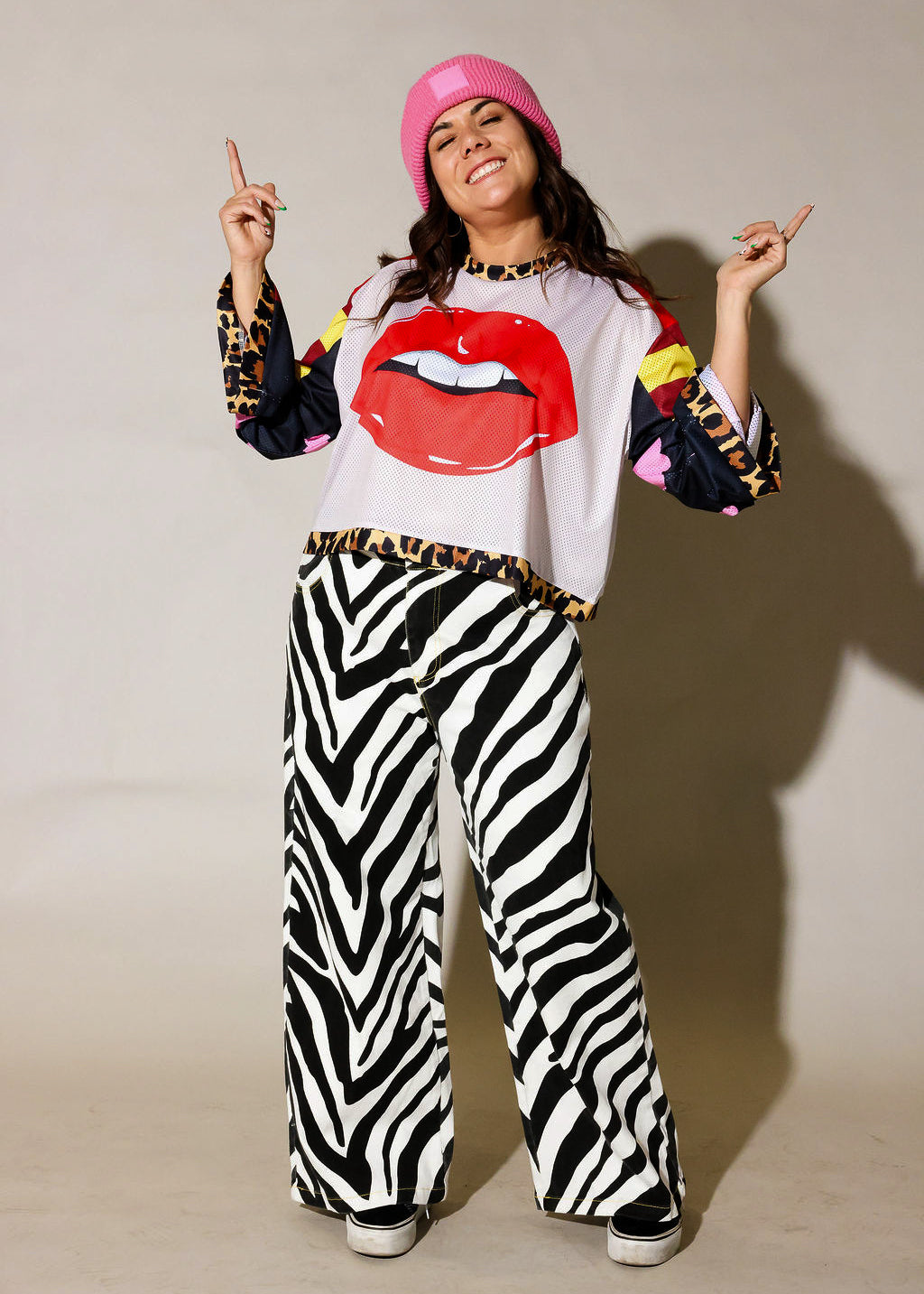 Person wearing a colorful outfit with zebra print pants, a white shirt with red lips design, and a pink hat, frontal view