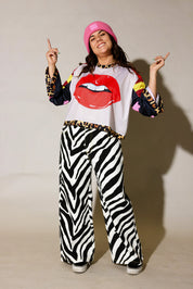 Person wearing a colorful outfit with zebra print pants, a white shirt with red lips design, and a pink hat, frontal view