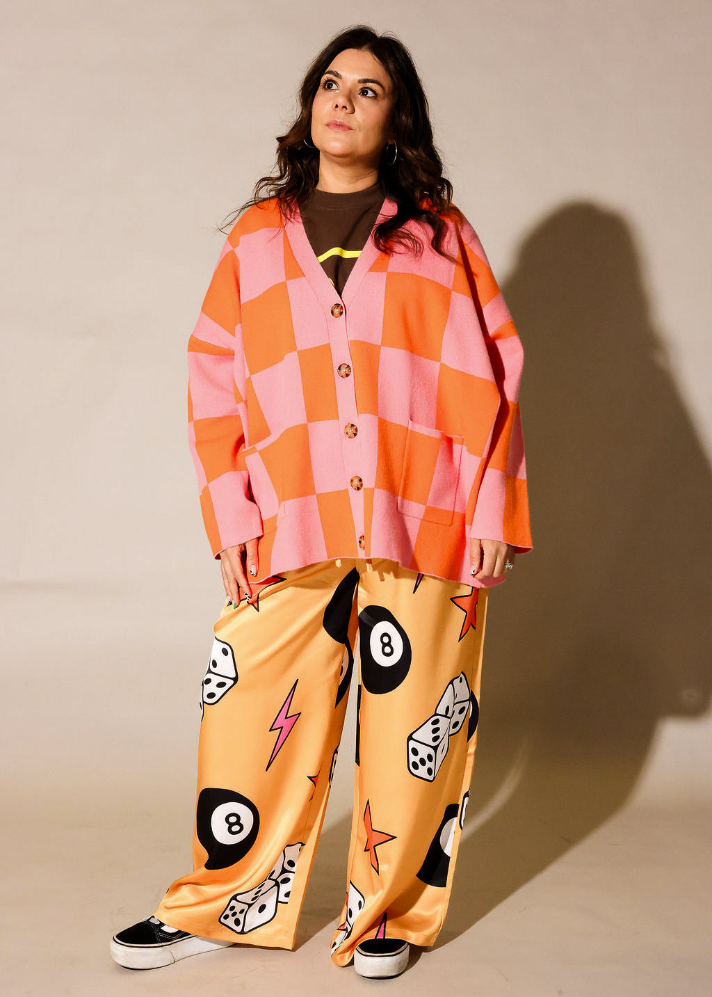 A woman modeling a matching two-piece lounge set featuring a pink and orange checkered button-up shirt and yellow pants with black-and-white billiards-themed graphics, set against a plain beige studio background, frontal view