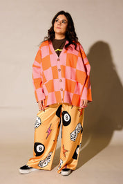 A woman modeling a matching two-piece lounge set featuring a pink and orange checkered button-up shirt and yellow pants with black-and-white billiards-themed graphics, set against a plain beige studio background, frontal view
