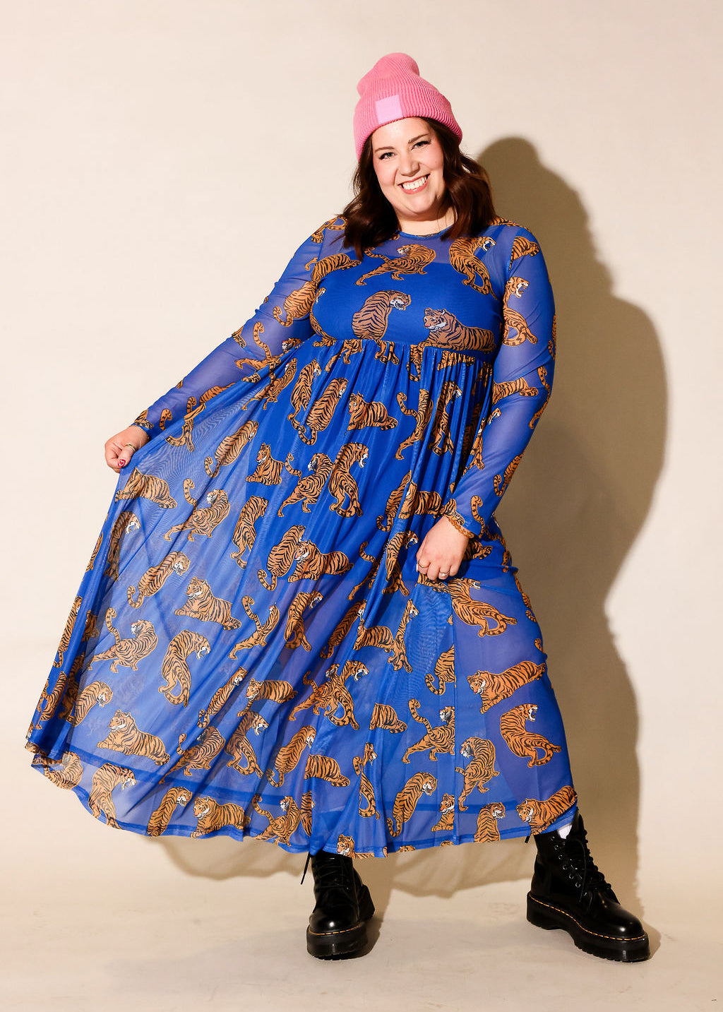 Model wearing a long sleeve blue mesh dress with tiger patterns and a pink beanie, and black boots, against a plain background, frontal view