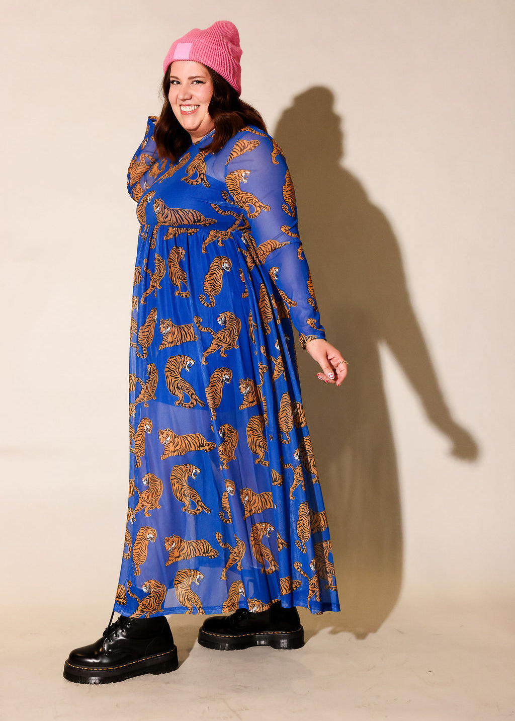 Model wearing a long sleeve blue mesh dress with tiger patterns and a pink beanie, and black boots, against a plain background, side view