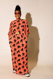 Model wearing an orange plisse short sleeve and skirt set with black animal prints, black beanie, and black and white boots, on a beige background, frontal view