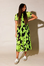 A woman modeling a short-sleeve midi dress with a black and neon yellow-green cow print, paired with white sneakers, against a plain beige studio background, frontal view