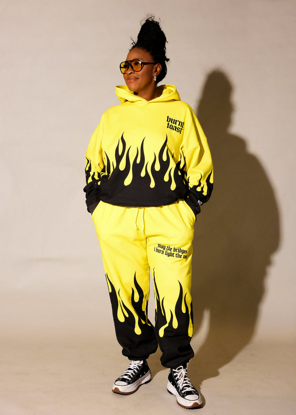 Model wearing a yellow hoodie and jogger set  with black flame design and 'burnt toast' logo, standing in front of a beige background, frontal view