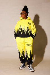 Model wearing a yellow hoodie and jogger set  with black flame design and 'burnt toast' logo, standing in front of a beige background, frontal view