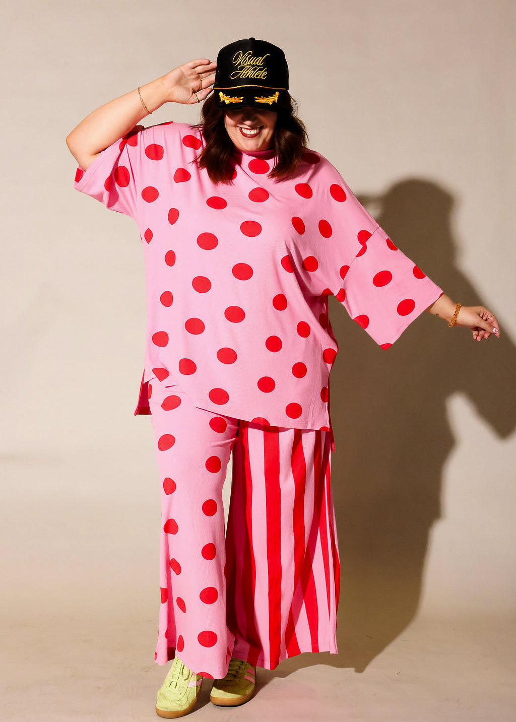 Model wearing a pink short sleeve set with red polka dots and striped pants, holding a black cap with yellow text, frontal view