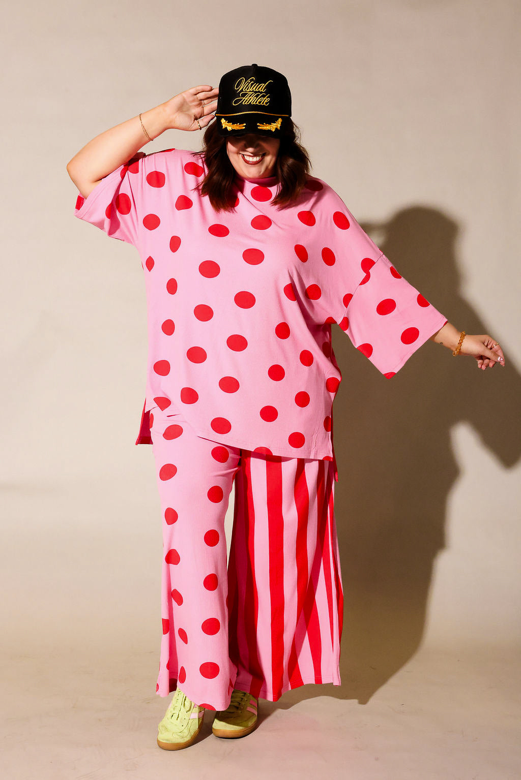 Model wearing a pink short sleeve set with red polka dots and striped pants, holding a black cap with yellow text, frontal view