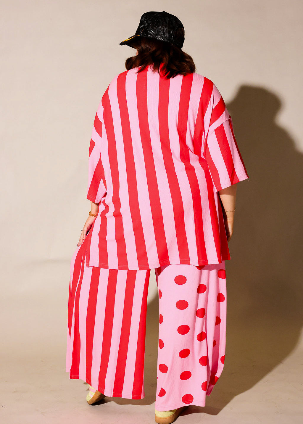 Model wearing a red and white striped set with a black trucker at on a beige background, back view